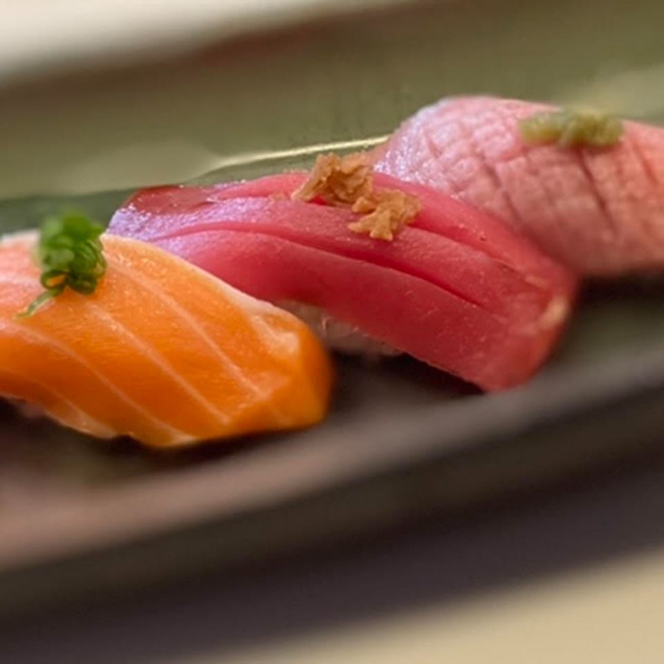 Three pieces of sushi, salmon, tuna, and seared tuna, garnished with herbs on a dark plate.
